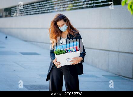 Depressed business woman with face mask outside office with personal staff box feeling hopeless after being fired from work. Coronavirus job losses, l Stock Photo