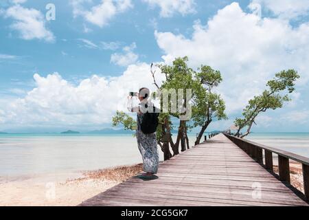 Tourist woman using mobile phone taking picture of ocean on the wooden bridge. Stock Photo