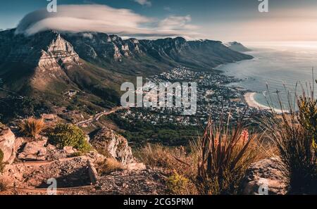 Cloud flows over Table Mountain, with Camps Bay beach visible during sunset hike up Lion's Head. Stock Photo