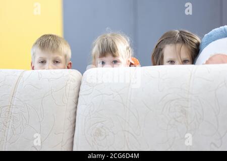 Boy and two girls hide behind sofa and look out. Stock Photo