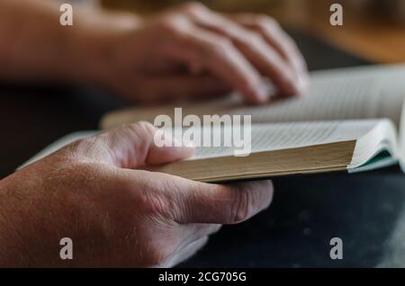 Male hands are holding an open book. A mature man is reading a thick hardback book on a black table. Selective focus. Stock Photo