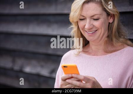 Outdoor portrait of an attractive middle aged blonde woman smiling using mobile cell phone for text messaging or social media Stock Photo
