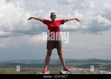 An adult woman stands, arms outstretched, on a high mountain, against the backdrop of a plain and a cloudy sky. Stock Photo
