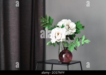 Two white peony flowers in glass vase on coffee table near window with curtain on dark background of home interior. Copy space dark mood Stock Photo