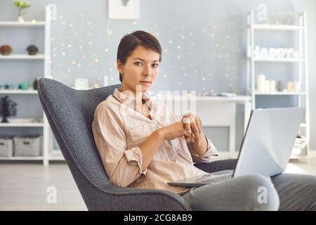 Serious businesswoman sitting in comfortable armchair with laptop in cozy home office Stock Photo