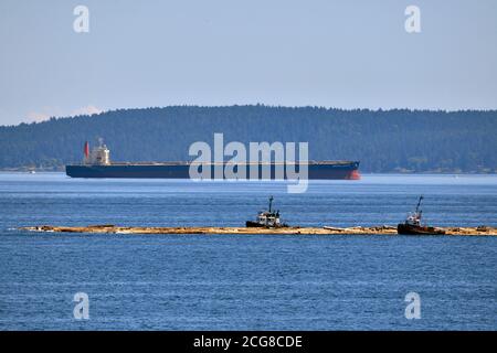 Two tug boats working a log boom in front of an ocean going ship anchored in the Stuart channel near Vancouver Island British Columbia Canada. Stock Photo