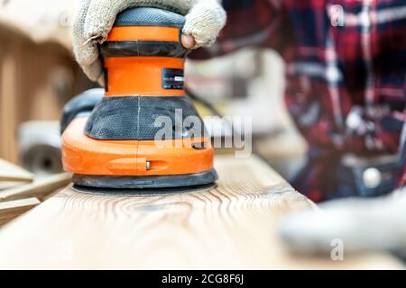 Closeup professional carpenter hand grinding raw wood plank with orbital sander electric machine in carpentry diy workshop. Detail of furniture Stock Photo