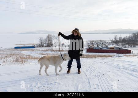 A young man walks with a big dog in the countryside against a frozen lake in winter. Stock Photo