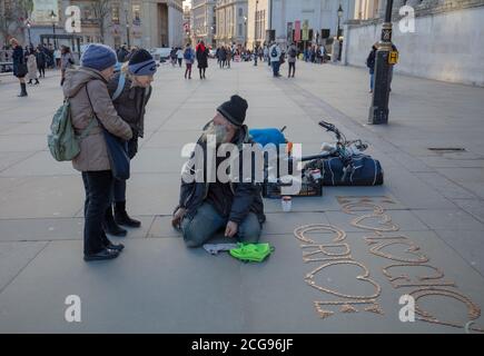 A busker, who made a design with copper coins on the pavement of Trafalgar Square, seen having a talk with two women, all three being senior citizens. Stock Photo