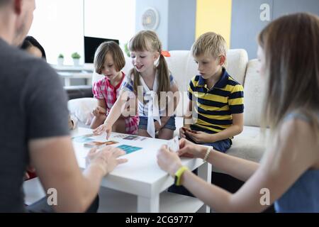 Adults and children are sitting around table on which playing cards are located girl reaches for card with her hand. Stock Photo
