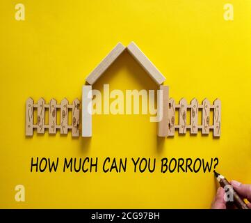 Hand writing 'how much can you borrow' on beautiful yellow background. Model of a wooden house. Male hand, wooden fence. Copy space. Business concept. Stock Photo