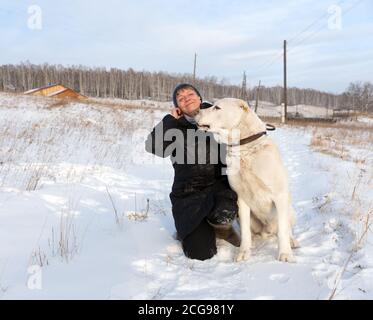 A happy woman is sitting with a Central Asian Shepherd on a winter rural road against the backdrop of the village. Stock Photo