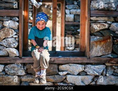 Young Sherpa Girl with Bandana Headband Sits on the window frame of under construction house and smiles at the camera in remote Manaslu region of Nepa Stock Photo