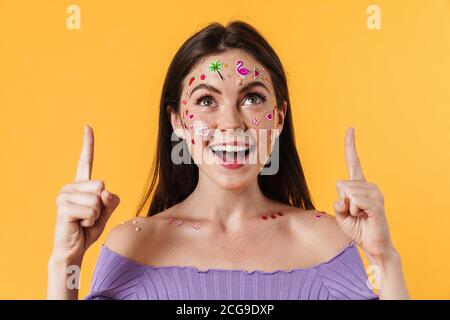 Image of smiling woman with stickers on face pointing fingers upward isolated over yellow wall Stock Photo