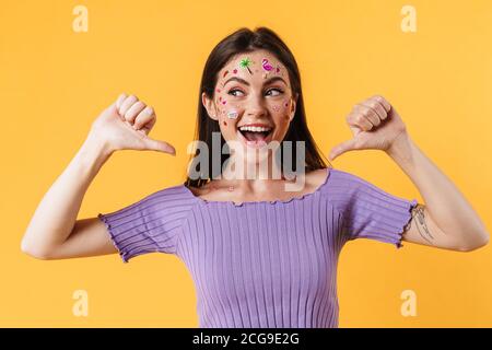 Image of excited woman with stickers on face pointing fingers at herself isolated over yellow wall Stock Photo