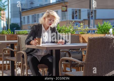 young woman in a black jacket and glasses sitting at a table in a street cafe, looking through documents. Business activity, conclusion of contract Stock Photo