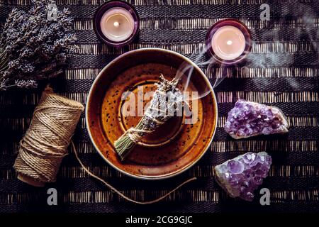Homemade herbal lavender (lavendula) smudge stick smoldering on brown plate with candles and amethyst crystal clusters for decoration. Spiritual home Stock Photo