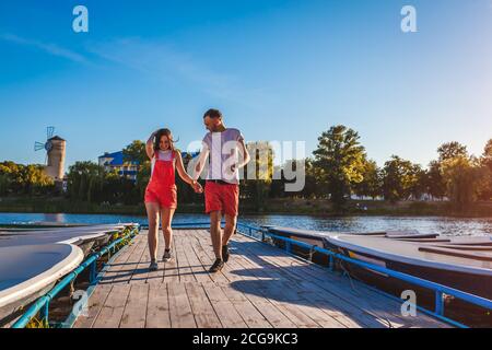 Young man and woman running along summer river dock. Couple having fun at sunset holding hands. People relaxing Stock Photo