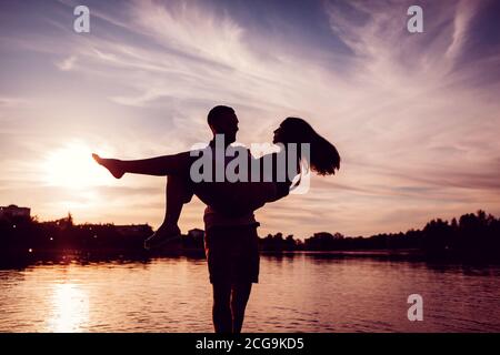 Man holding woman in hands. Couple having fun on river bank at sunset. Silhouettes of people Stock Photo