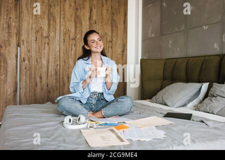 Image of a young smiling positive girl indoors at home drinking coffee while sitting near paper documents Stock Photo