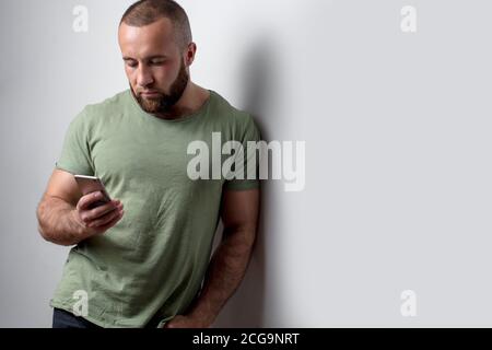 Bearded fashionable guy with masculine appearance downloads photos for blog via modern cell phone, has serious pensive expression, leaning on white wa Stock Photo