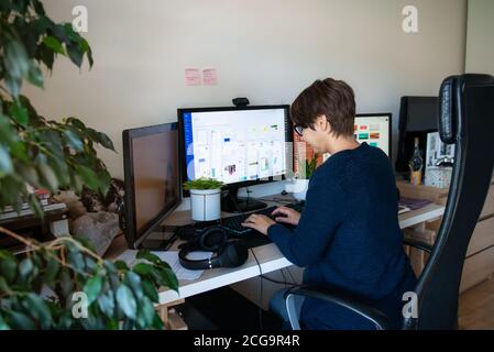 Back view woman in glasses typing on pc with headsets, few computers on her work desk at home working space. Remote work concept. Freelancer workspace Stock Photo