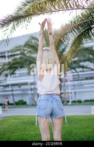 Young female runner in summer active clothes stretching arms before running at the sunny morning standing near big palm in hotel garden Stock Photo