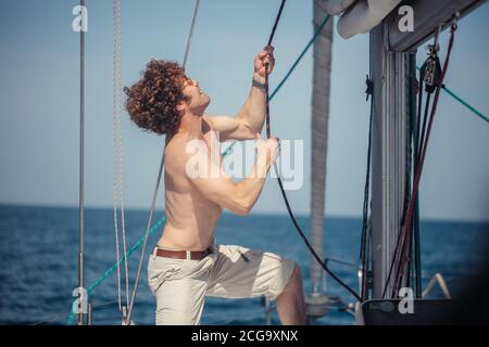 Young handsome hipster sailor man with fuzzy hairs sailing a boat to take his friends to a sea trip, pulling the rope Stock Photo