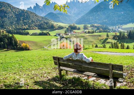 Woman sitting on bench and enjoying the view village of Santa Magdalena on the slopes of the Dolomites in Italy in the valley of Val di Funes. Stock Photo
