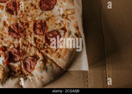 A large round juicy mouth-watering pizza with cheese and salami lies on a cardboard box from a delivery package. paper backing. close view. see flour. Stock Photo