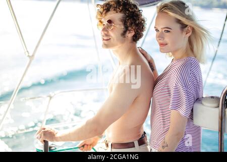Beautiful couple of lovers sailing on a yacht - Young attractive man holding rudder of a yacht and looking far away. Stock Photo