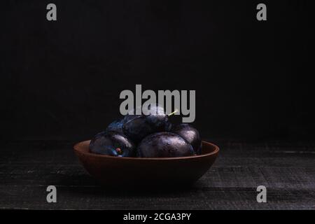 Fresh ripe plum fruits with water drops in wooden bowl on dark wood background, angle view Stock Photo