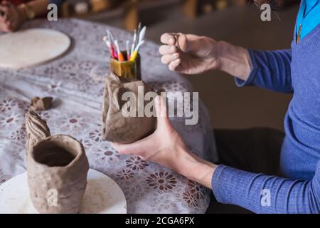 Professional male potter shaping mug in shape of human face with special tool in pottery workshop, studio. Crafting, artwork and handmade concept Stock Photo