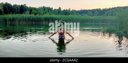 A girl in a swimsuit and with a bouquet of flowers on her head, stands in the water on the holiday of Ivan Kupala Stock Photo