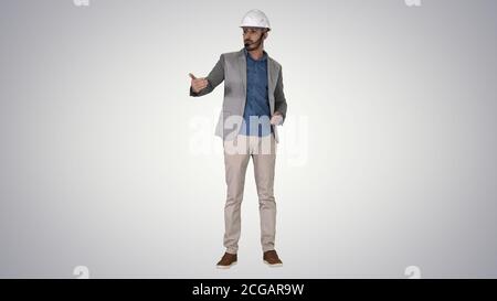 Architect man in helmet and head set talking to camera and making gestures showing on some objects on gradient background. Stock Photo