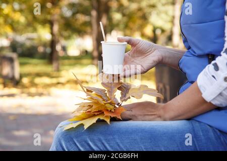 Young woman sitting in the city park and holding a cup of coffee and dry autumn leaves in her hands. Autumn composition. Stock Photo
