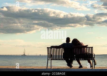 A young African American couple is sitting on a bench looking at sea and Chesapeake Bay bridge. Man  puts his arm over the shoulder of the woman w Stock Photo