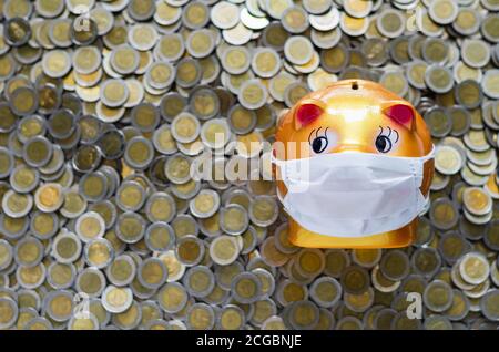 Piggy bank wearing white face mask on blurred background of Thai baht coins. Money saving concept in time of coronavirus pandemic. Stock Photo