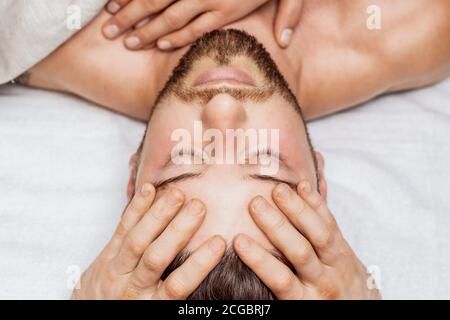 Handsome young man receiving relaxing head massage by four hand of two masseurs in health spa center Stock Photo