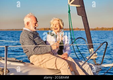 Having fun. Happy beautiful senior family couple drinking wine or champagne and laughing while relaxing on a sailboat or yacht deck floating in a calm Stock Photo