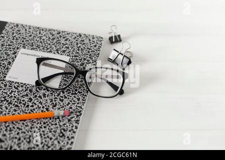 Black framed eyeglasses, wood pencil, black and white composition book laying on white wooden table surface with a pair black clips. Stock Photo