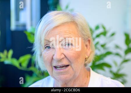 Candid portrait of an attractive 80 year old woman speaking to a friend with her eyes on the camera in close up against a green plant Stock Photo