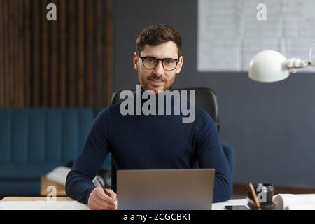 Young businessman working in office with laptop. Business portrait of handsome bearded man wearing eyeglasses sitting at workplace. Confident Stock Photo