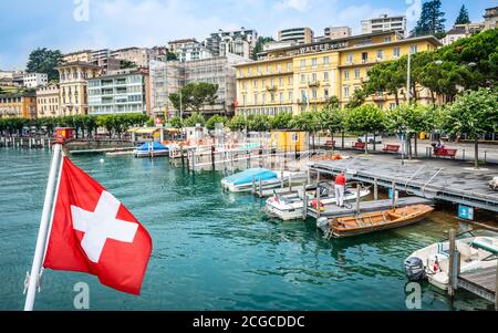 Lugano Switzerland , 1 July 2020 : Swiss flag and Lugano cityscape with boats on the shore of Lake Lugano Ticino Switzerland Stock Photo