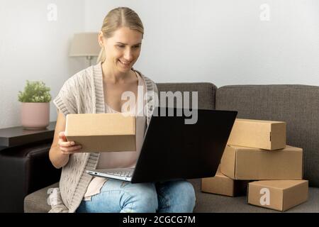 online shopping or small internet store business concept - young smiling woman sitting on couch with laptop and cardboard boxes Stock Photo