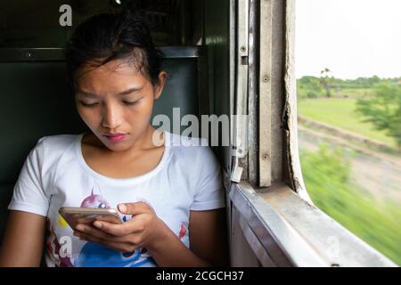 A young woman with mobile phone rides in old train. Asian girl with smartphone sits at retro wagon, Thailand. Stock Photo