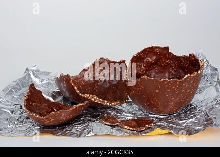 Broken chocolate Easter egg with chopped hazelnuts on a foil sheet against a white background, UK. Stock Photo