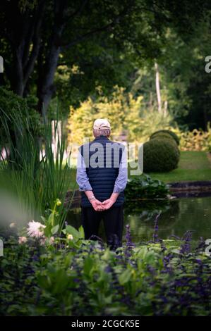 A senior man standing and looking out over a lush garden and pond Stock Photo