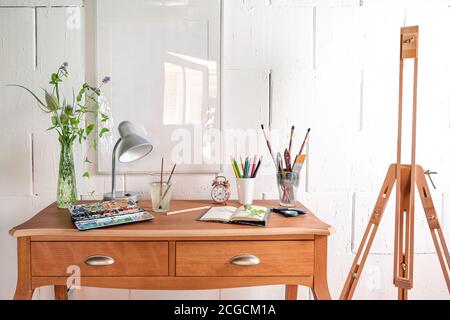 Creative place for art work or hobby, crayons, sketchbook, watercolor and brushes on a small wooden desk and an empty easel against a white painted wa Stock Photo