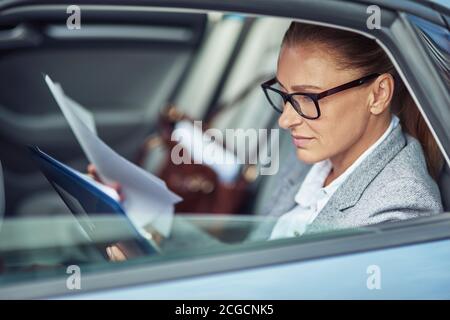 Side view of a focused middle aged business woman wearing eyeglasses analyzing documents while sitting on back seat in the car Stock Photo